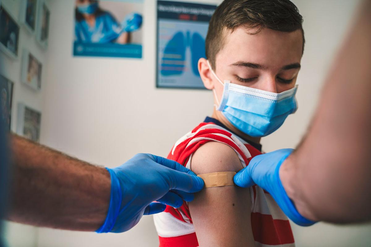 Masked child watching as a health care worker applies an adhesive bandage
