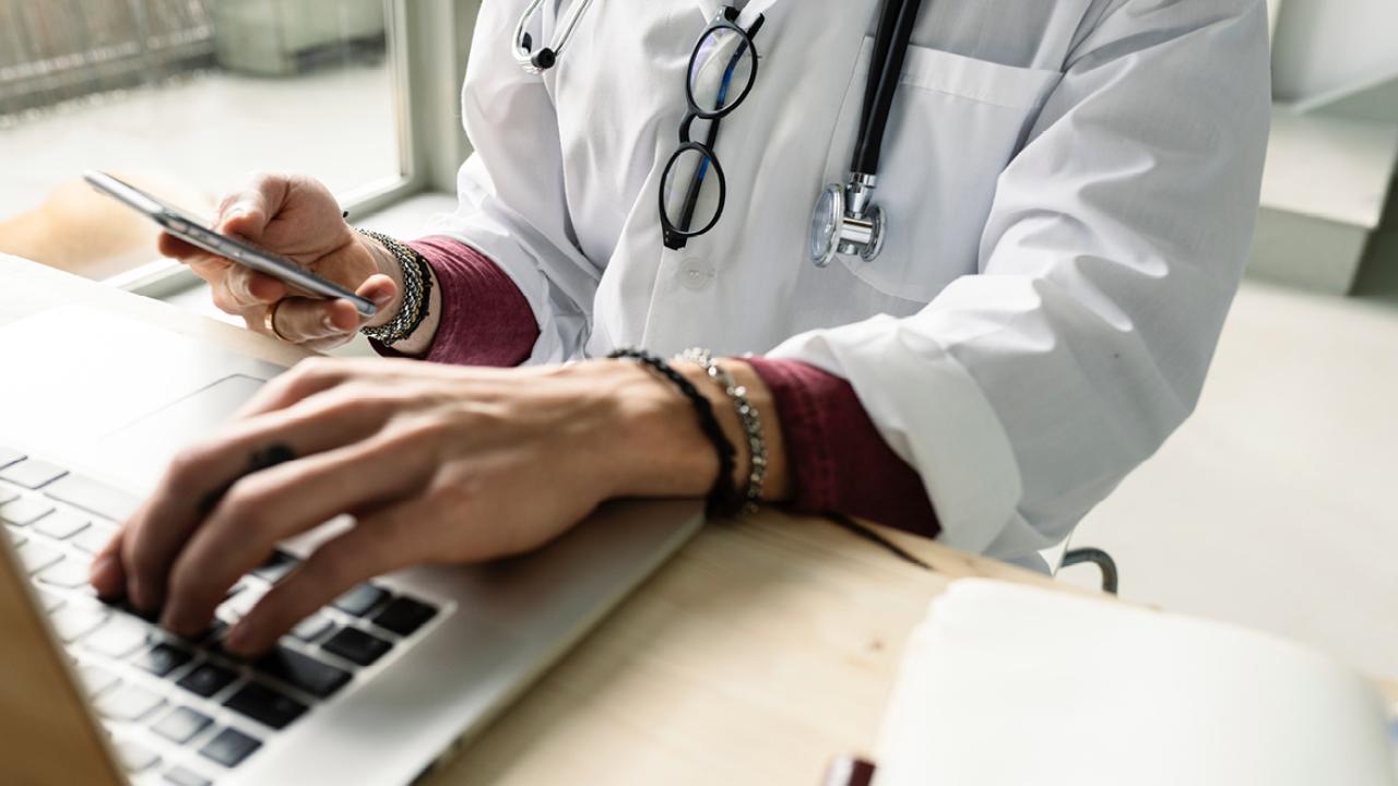 Doctor typing on laptop at desk with stethoscope around their neck