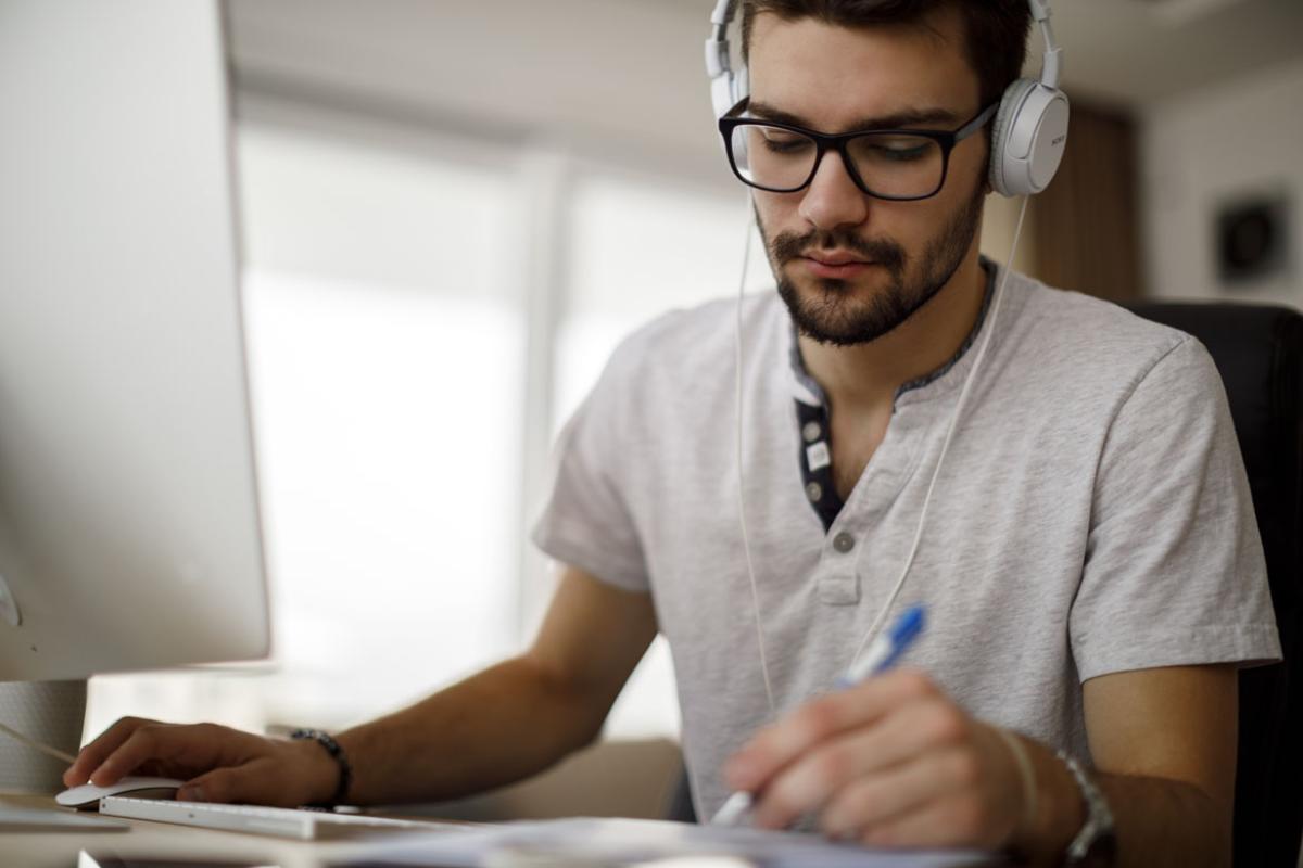 Man wearing headphones working at a computer, taking notes.