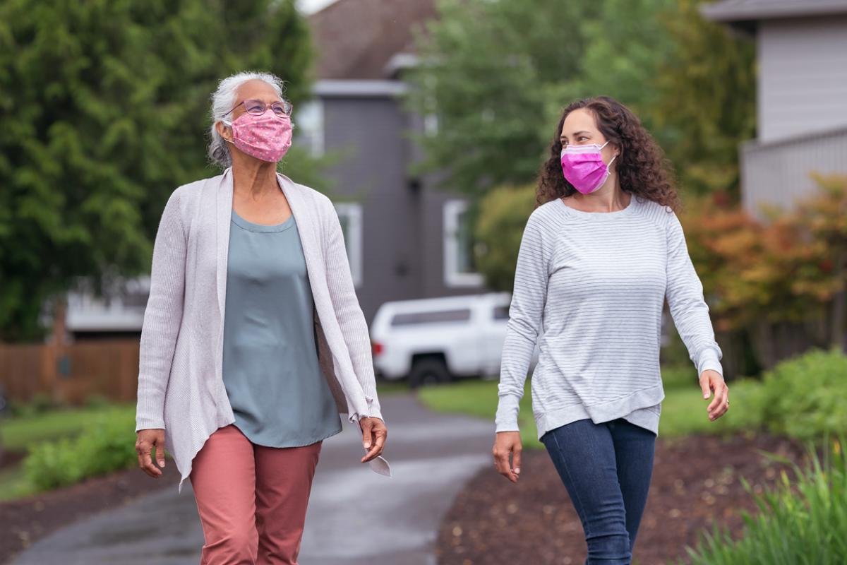 An older woman and a younger woman, both wearing masks, walking outside in a suburban neighborhood.
