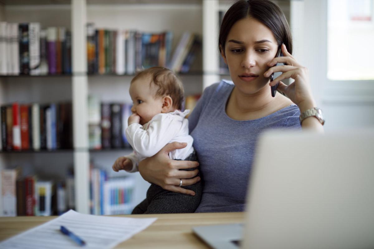 Seated woman talking on a cell phone while looking at a computer and holding a baby.