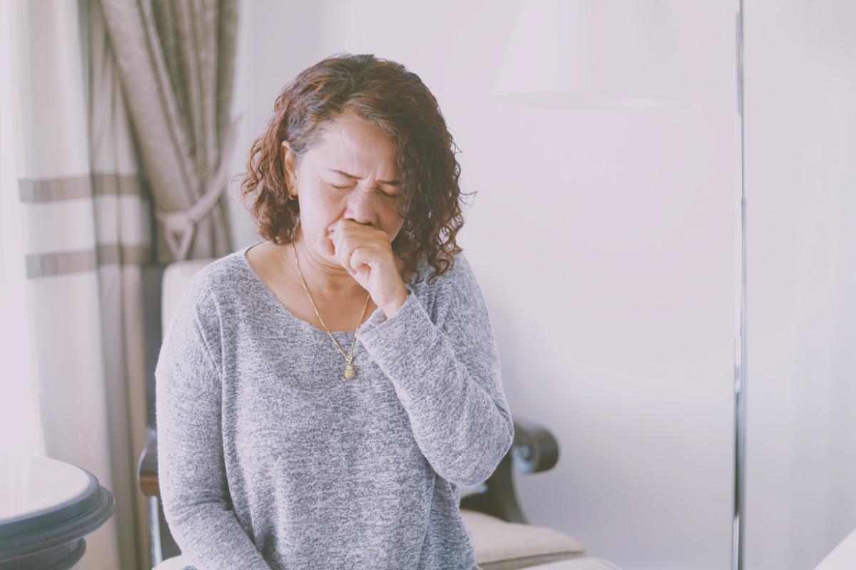 Woman in a white room coughing, covering her mouth.