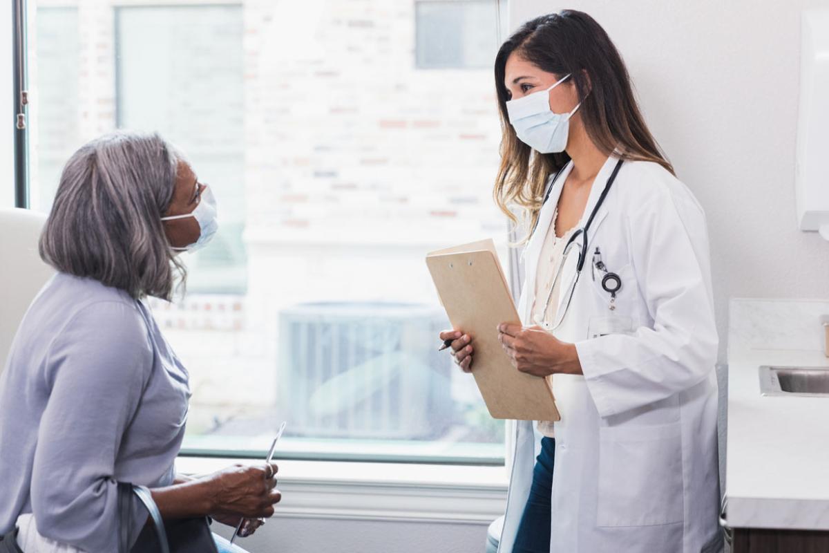 A patient and doctor talking in a white room both wearing masks.