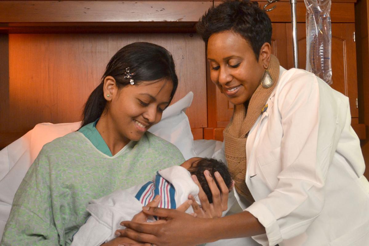 Veronica Gillispie-Bell, MD handing a new mother in a hospital bed her newborn.