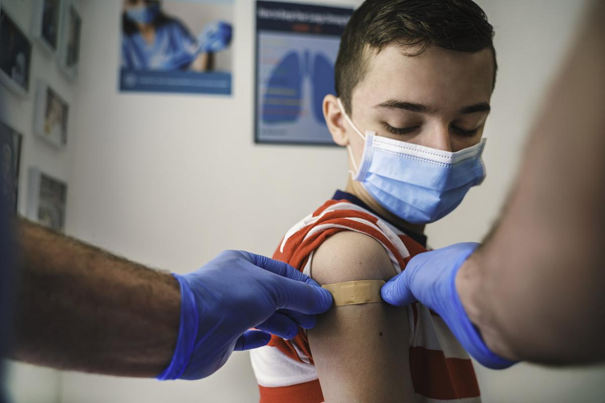 Young boy with mask looks on as a health professional applies an adhesive bandage to his right arm after getting a vaccination