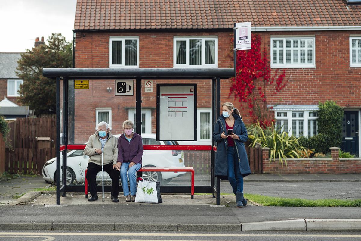 Three people, including an elderly couple, wearing masks and waiting at a bus stop shelter for a bus