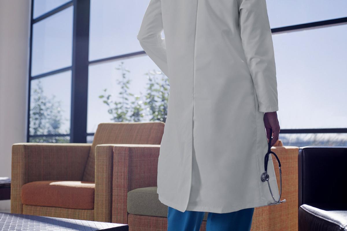 Shot from the back of a physician standing in a hospital waiting area looking out a set of windows, holding a stethoscope in the right hand