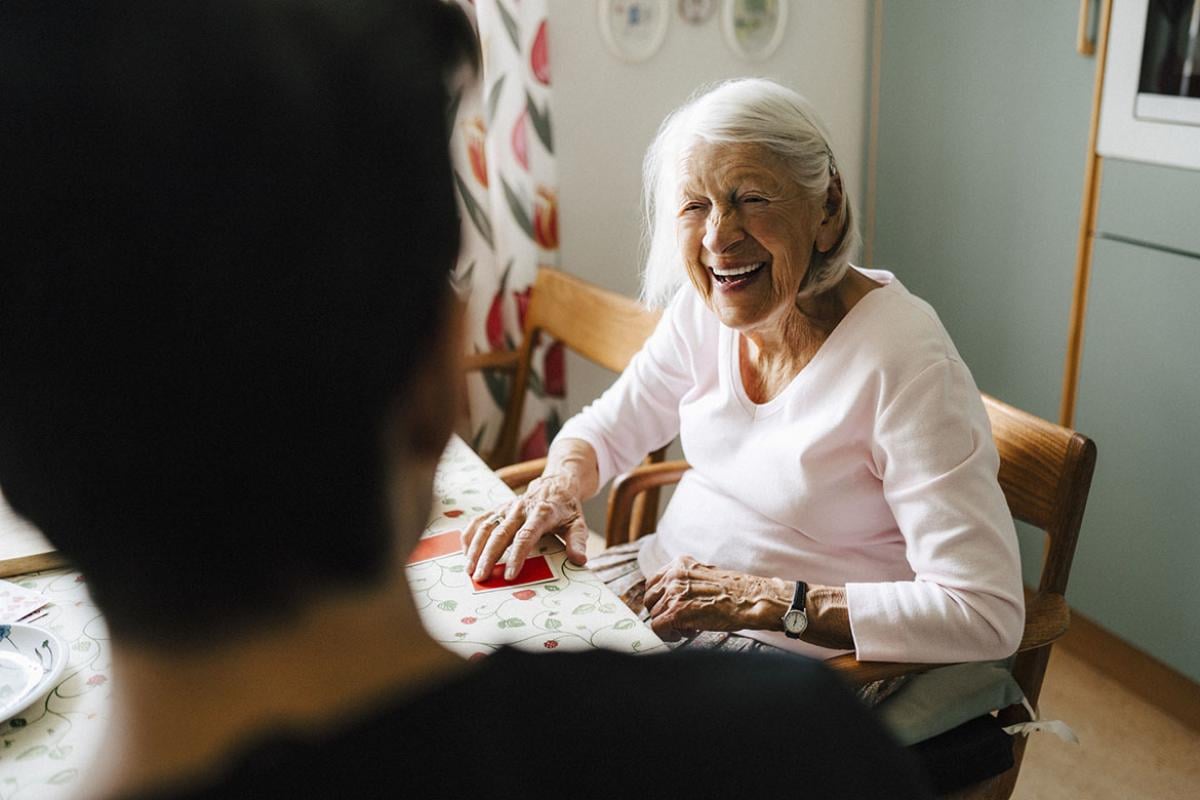 Smiling senior woman talking with caregiver while playing cards in kitchen