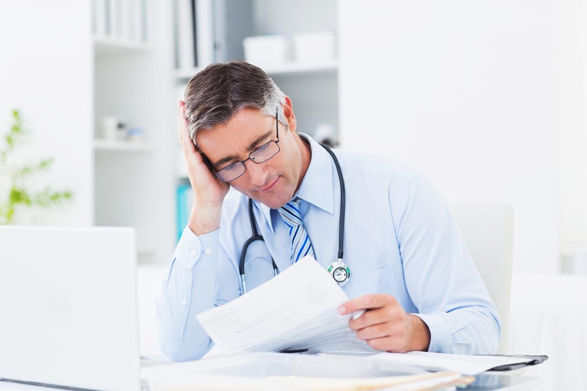 Physician resting hand on head reviewing paperwork. 