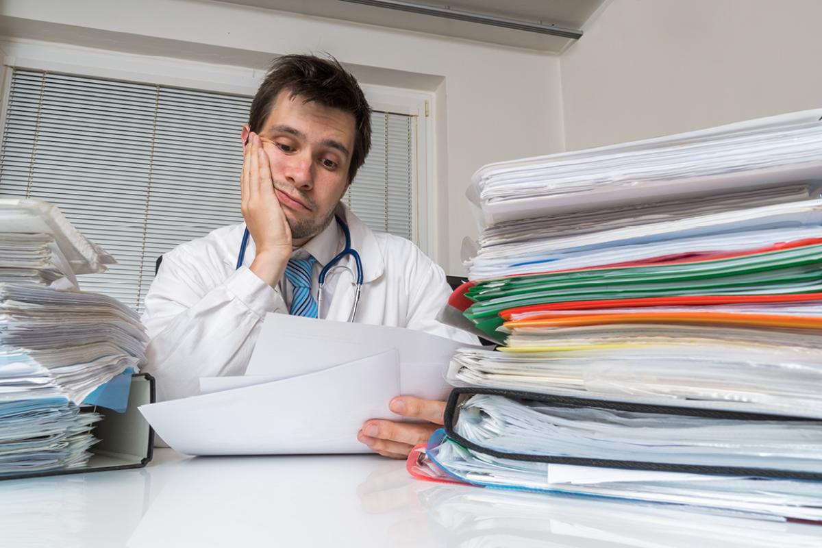 Dejected physician sits in front of a stack of paperwork. 