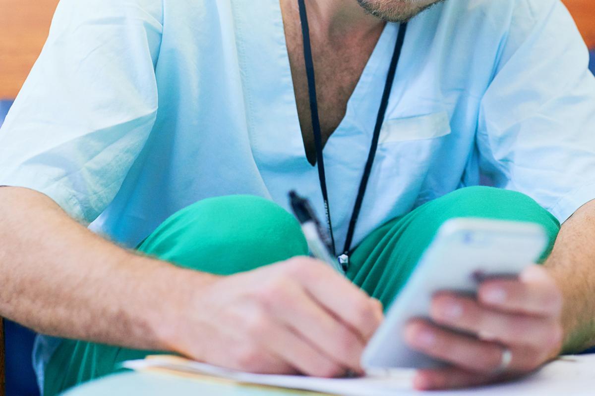 A doctor writing on a notepad while reading something off of a smartphone.